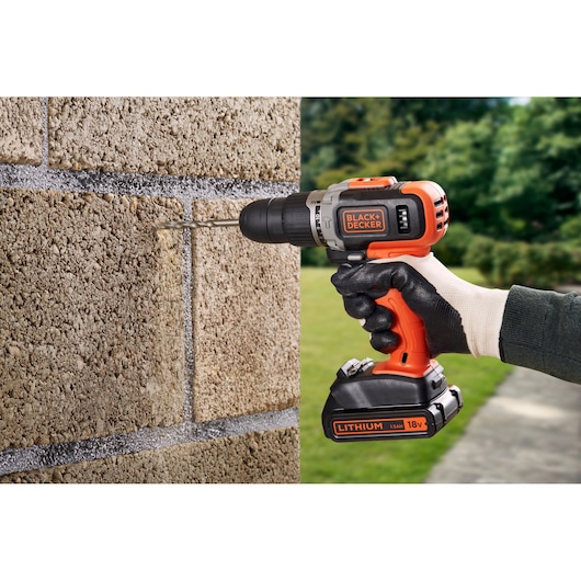 A person wearing gloves uses a BLACK+DECKER cordless drill to drill into a concrete block wall outdoors. The drill is orange and black and labeled with 'BLACK+DECKER' and 'LITHIUM 18V.'