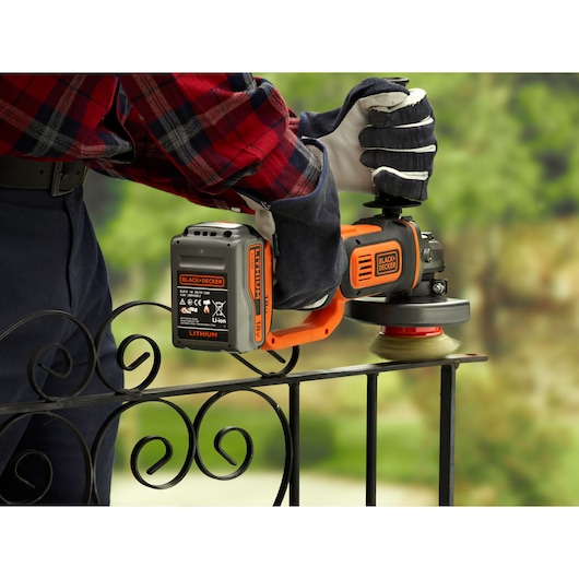 A close-up view of gloved hands using a BLACK+DECKER cordless grinder on a metal railing outdoors, with the focus on the tool's battery and brush attachment.