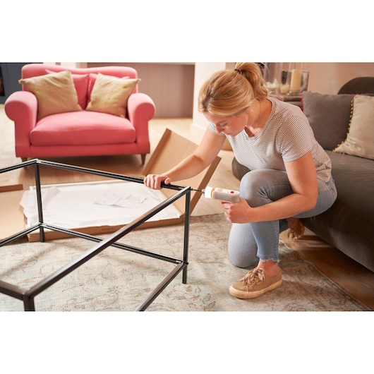 A person kneeling on the floor is using a BLACK+DECKER cordless screwdriver to assemble a metal-framed table in a living room. There is a pink couch with pillows and an open cardboard box with assembly instructions in the background.