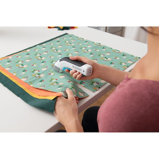 Photo of BLACK+DECKER BCSC115 electric scissors held cutting floral fabric on a white table.