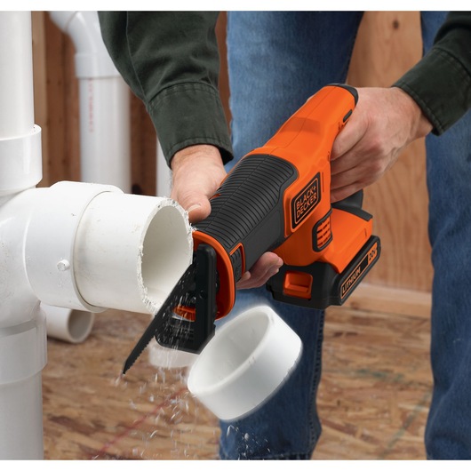 A person using a BLACK+DECKER cordless reciprocating saw to cut through a white PVC pipe. The saw is orange and black, with visible branding and a Lithium Ion battery.