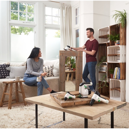 BLACKDECKER BDST60120 cordless power tools on coffee table in bright living room with two people in background.