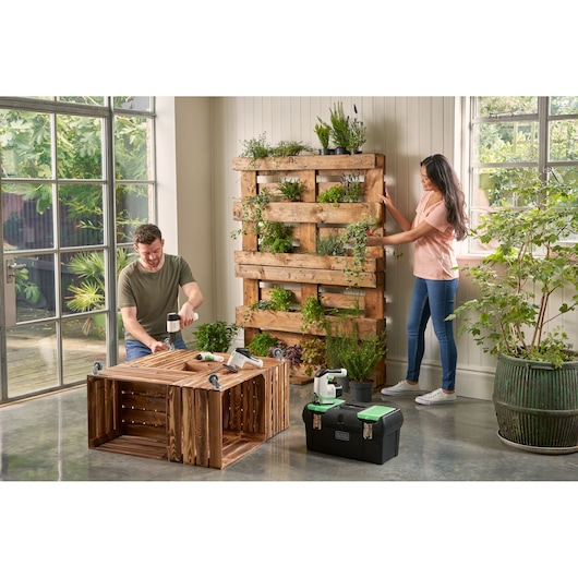 BLACKDECKER BDST60120 black toolbox with green latches on floor in indoor garden workspace, wood planters and plants visible.