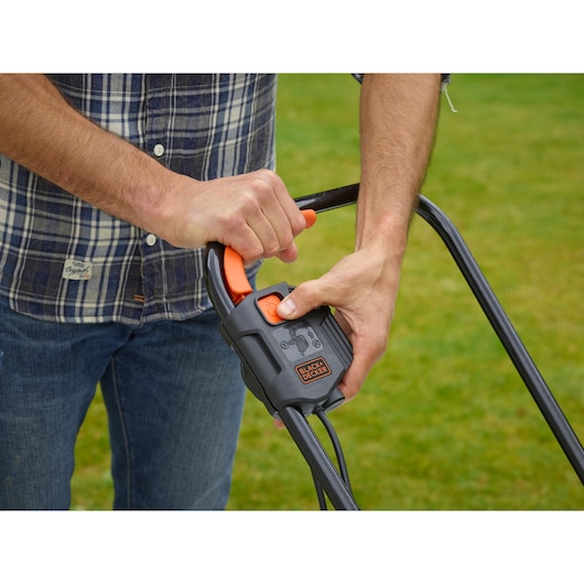 Close-up of hands operating BLACK+DECKER BEMW451 lawn mower handle with control lever on grass.