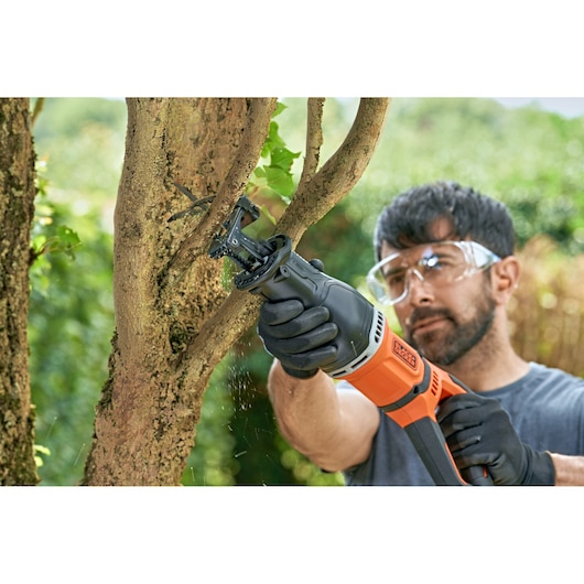 A person wearing gloves uses an orange and black reciprocating saw to cut a tree branch outdoors. The face of the person is blurred for privacy.