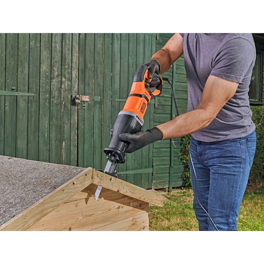 A person using a BLACK+DECKER BES301 reciprocating saw to cut a piece of wood outdoors, wearing gloves and holding the saw on a wooden structure.