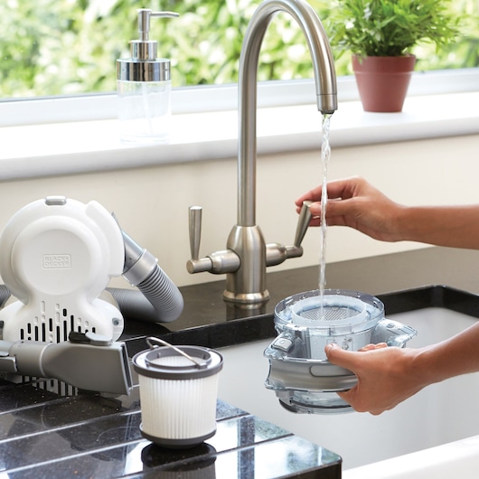 Photo of BLACK+DECKER PD1820L handheld vacuum parts being cleaned under a kitchen faucet, filter and canister visible.