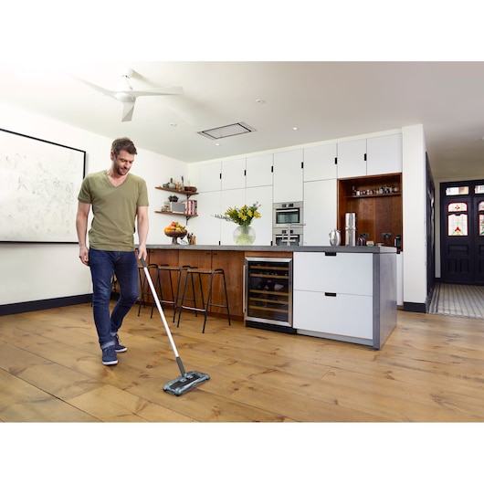 A person using a BLACK+DECKER floor cleaning tool to clean a wooden floor in a modern kitchen. The kitchen has white cabinets, a countertop with a vase of yellow flowers, and stools.