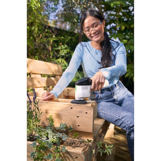 BLACK+DECKER REVDS12C cordless sander in use outdoors on wooden bench.